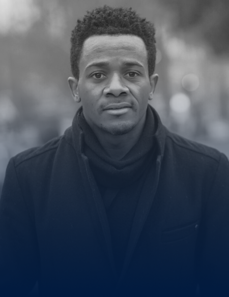 Portrait of a young man with short curly hair wearing a dark coat and scarf, looking directly at the camera outdoors.