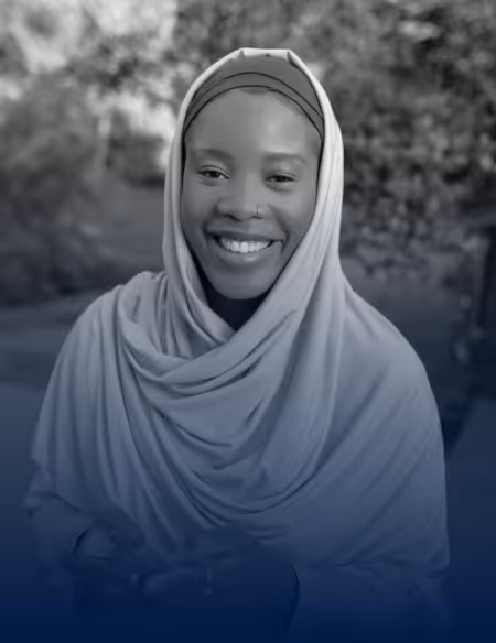 Smiling woman wearing a hijab, holding a phone, outdoors with blurred greenery in the background.