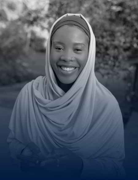 Smiling woman wearing a hijab, holding a phone, outdoors with blurred greenery in the background.