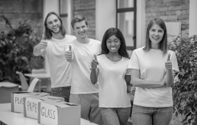 Four diverse young adults standing behind labeled recycling bins giving thumbs up.