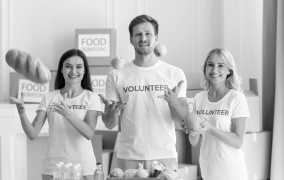 Three volunteers smiling and pointing at food donation boxes in a community center.
