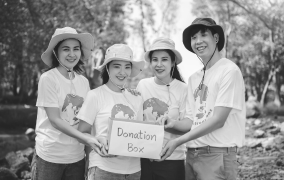 Four smiling volunteers wearing hats hold a donation box outdoors in a natural setting.