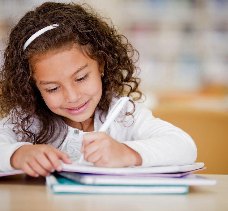 Smiling young girl with curly hair writing in a notebook at a desk.