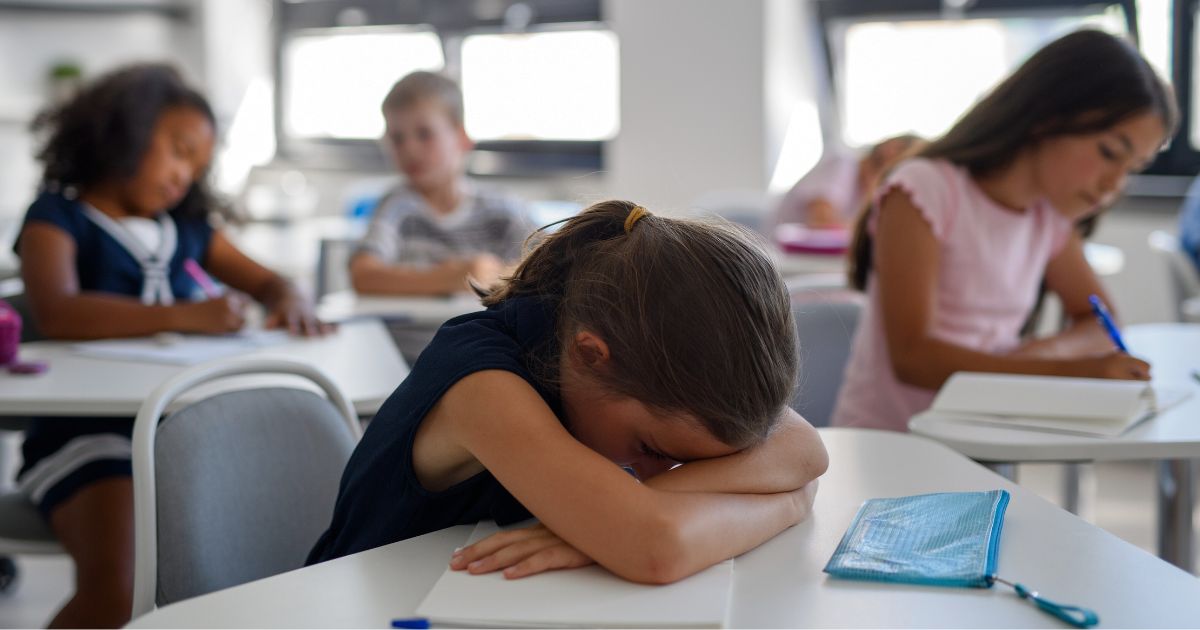 A young girl in a classroom rests her head on folded arms on her desk while other children are writing around her.
