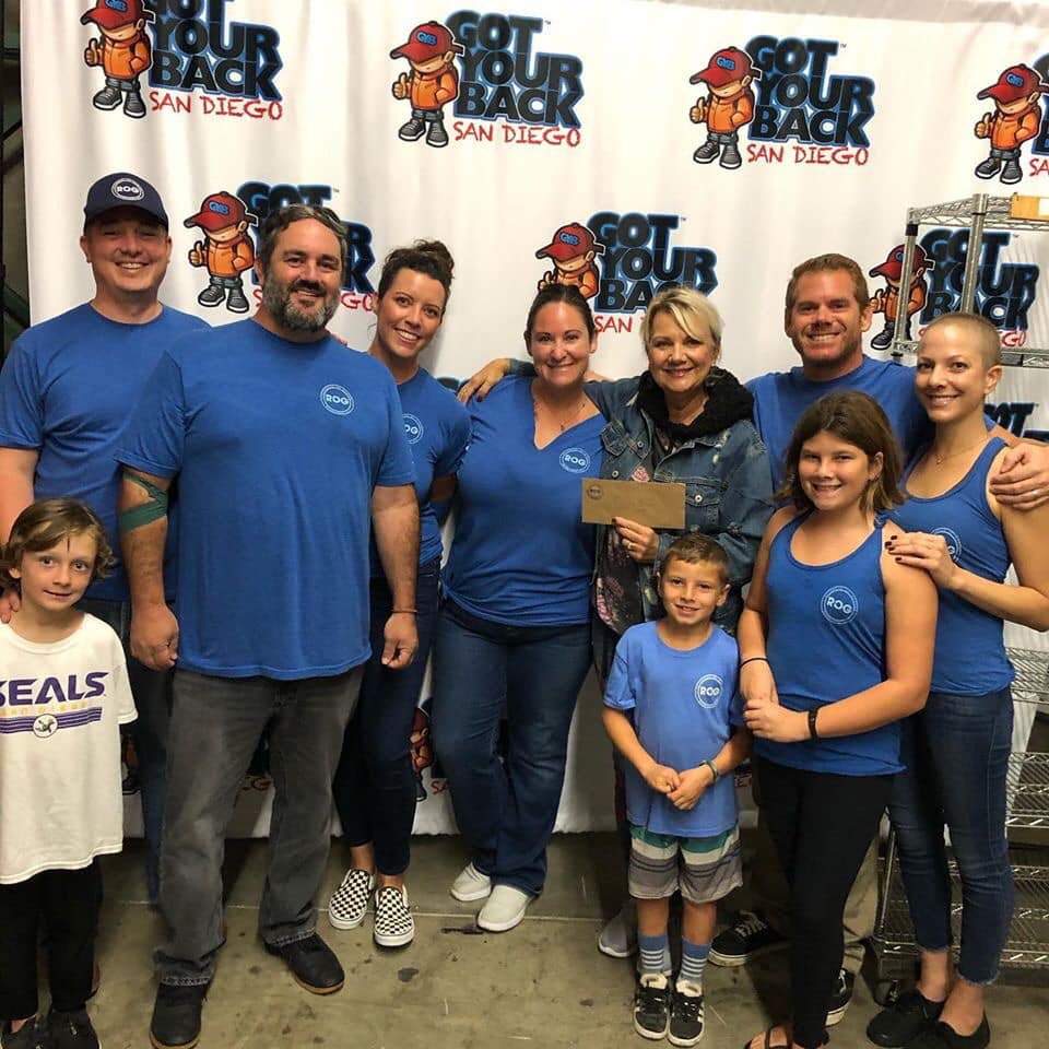 Group of adults and children smiling and posing in front of a Got Your Back San Diego backdrop, many wearing blue shirts with a circular logo.