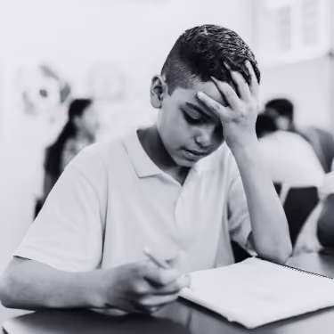 Boy concentrating with his hand on forehead while writing in a notebook in a classroom.