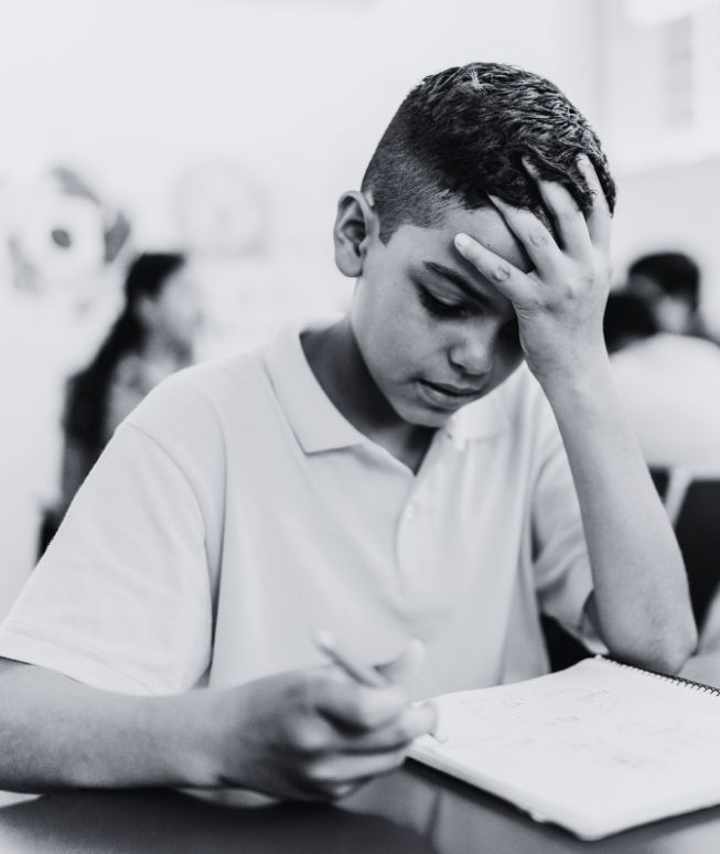 Boy concentrating with his hand on forehead while writing in a notebook in a classroom.