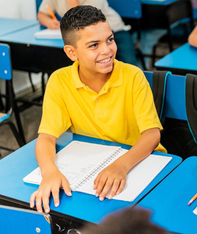Smiling boy in a yellow shirt sitting at a blue desk with an open notebook in a classroom.