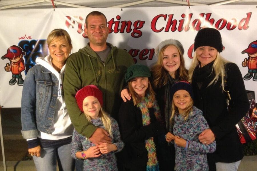 Group of three adults and four children standing and smiling in front of a banner that reads 'Fighting Childhood Cancer.'