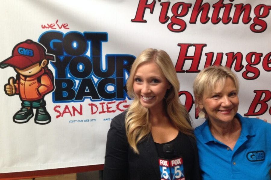 Two smiling women in front of a banner reading 'We've Got Your Back San Diego' with a cartoon character and partial text about fighting hunger; one woman holds a Fox 5 microphone.