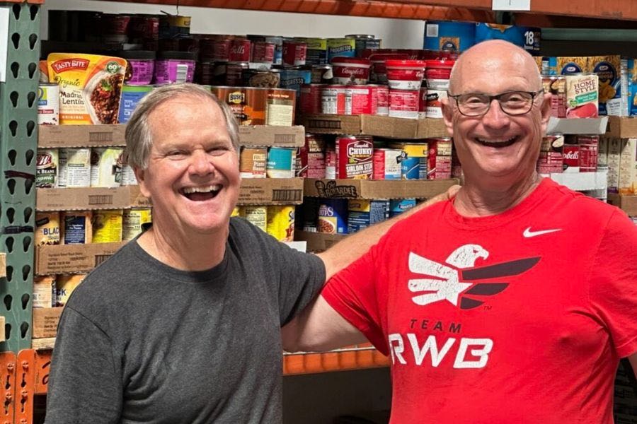 Two smiling men, one in a gray shirt and the other wearing glasses and a red Team RWB t-shirt, standing side by side in front of shelves stocked with canned goods.