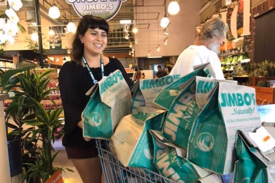 Woman pushing a shopping cart full of green and white Jimbo's grocery bags inside a store.