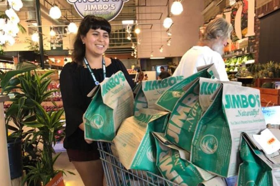Woman pushing a shopping cart full of green and white Jimbo's grocery bags inside a store.
