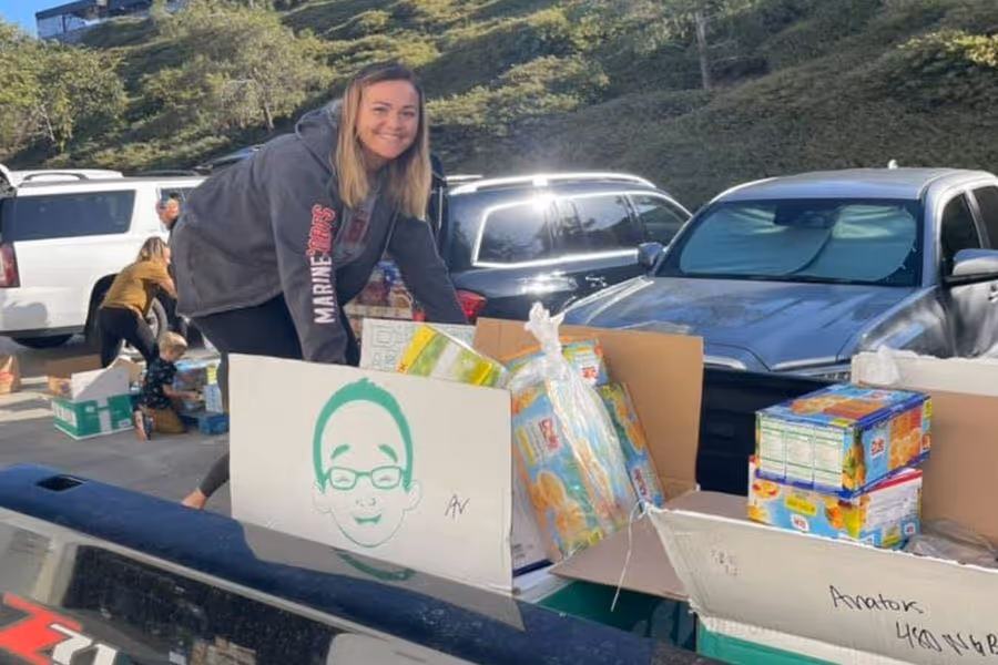 Woman smiling and organizing food boxes in the back of a vehicle during a food drive in a sunny outdoor parking area.