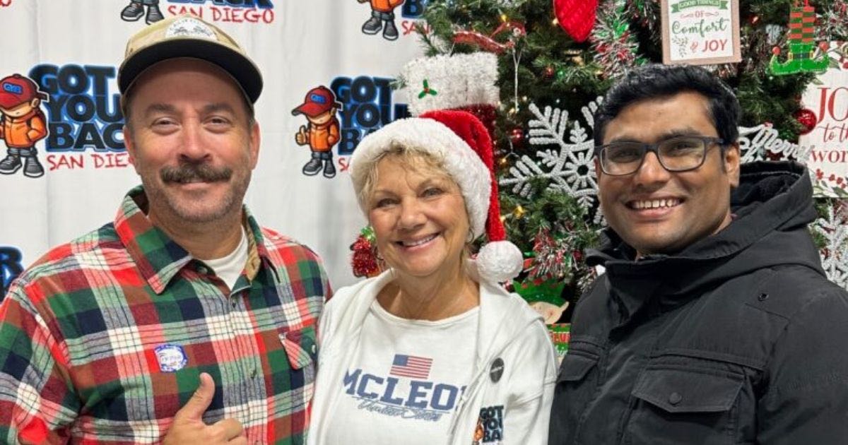 Three people smiling indoors in front of a Christmas tree and Got Your Back San Diego banner; woman in middle wears a Santa hat.