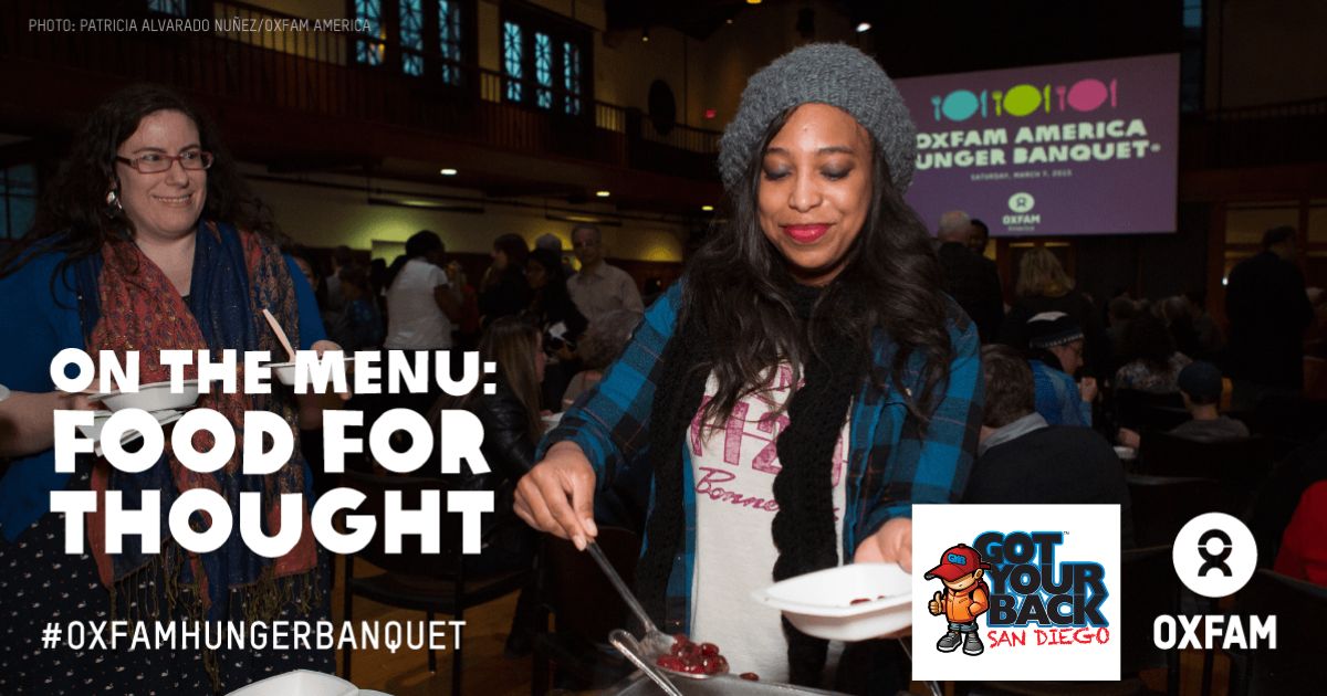 Two women serving and receiving food at an Oxfam America Hunger Banquet event with a screen in the background displaying the event name and date.