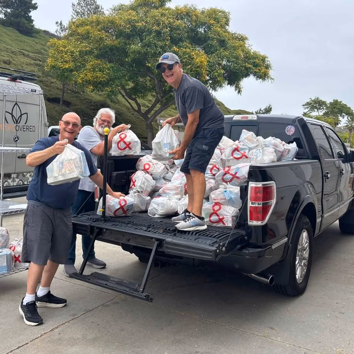 Three men unloading bags from a black pickup truck filled with white plastic bags outdoors.