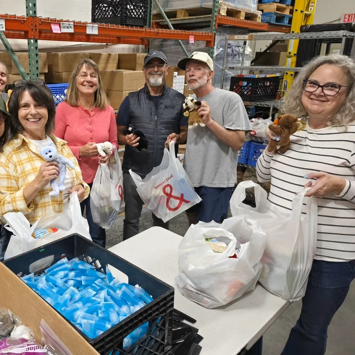 Group of smiling adults holding plastic bags and small stuffed animals in a warehouse setting.