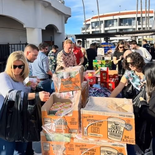 People gathering around tables with boxes of Cup Noodles and snacks outdoors on a sunny day.