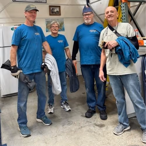 Four volunteers wearing casual clothes standing in a garage or workshop holding jackets and bags.