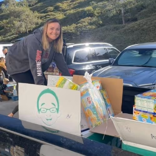 Woman smiling while organizing boxes of food and supplies outside near parked cars.