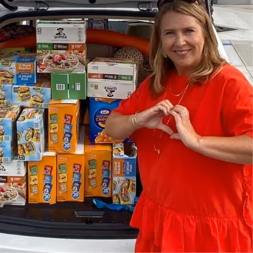 Woman in red shirt making a heart with hands beside car trunk filled with cereal boxes.