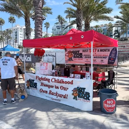 Outdoor charity booth with red canopy showing signs about fighting childhood hunger in San Diego.