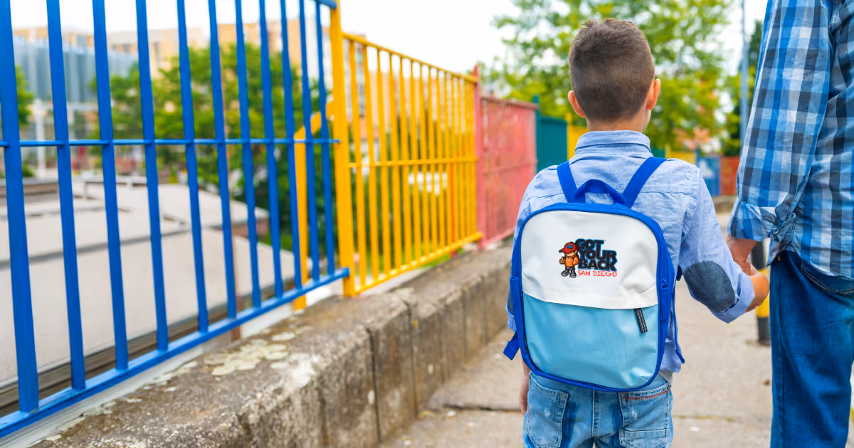 A young boy wearing a light blue shirt and backpack holding hands with an adult walking along a sidewalk next to a colorful fence in blue, yellow, and red.