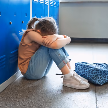 Young girl sitting on floor with head down and arms wrapped around knees next to blue lockers and backpack.