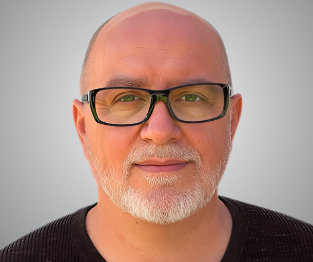 Porträt eines älteren Mannes mit Glatze, Brille und weißem Bart vor grauem Hintergrund.