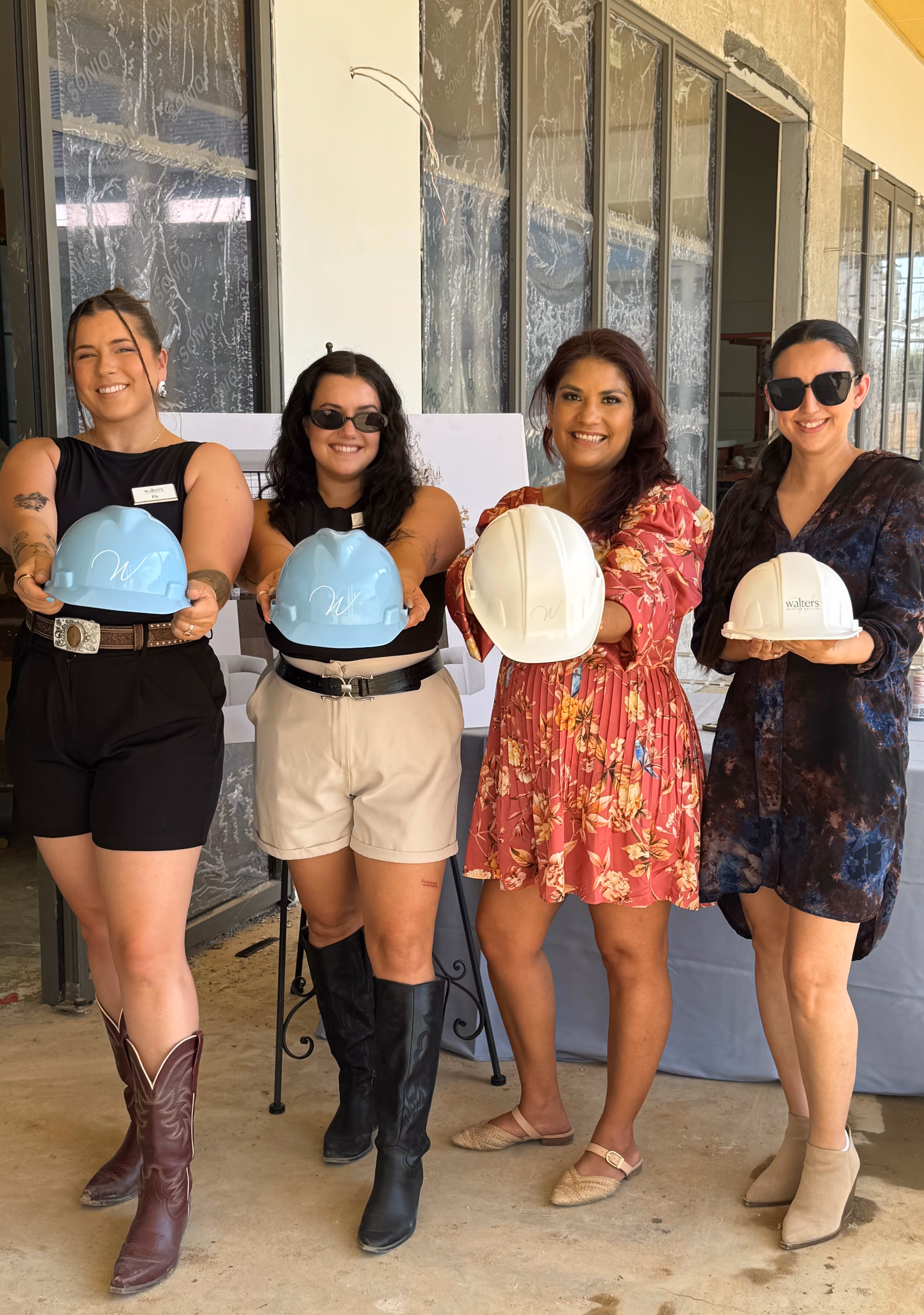 Four smiling women standing indoors holding blue and white construction helmets towards the camera.