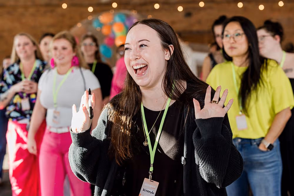 Smiling woman with dark hair and black sweater holding a pen and raising her hands, with a group of people in the background wearing name badges.