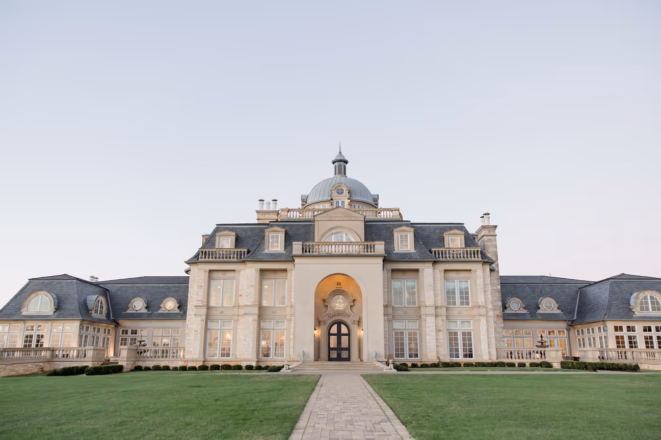 Large elegant mansion with stone facade, central dome, and symmetrical wings surrounded by green lawn.