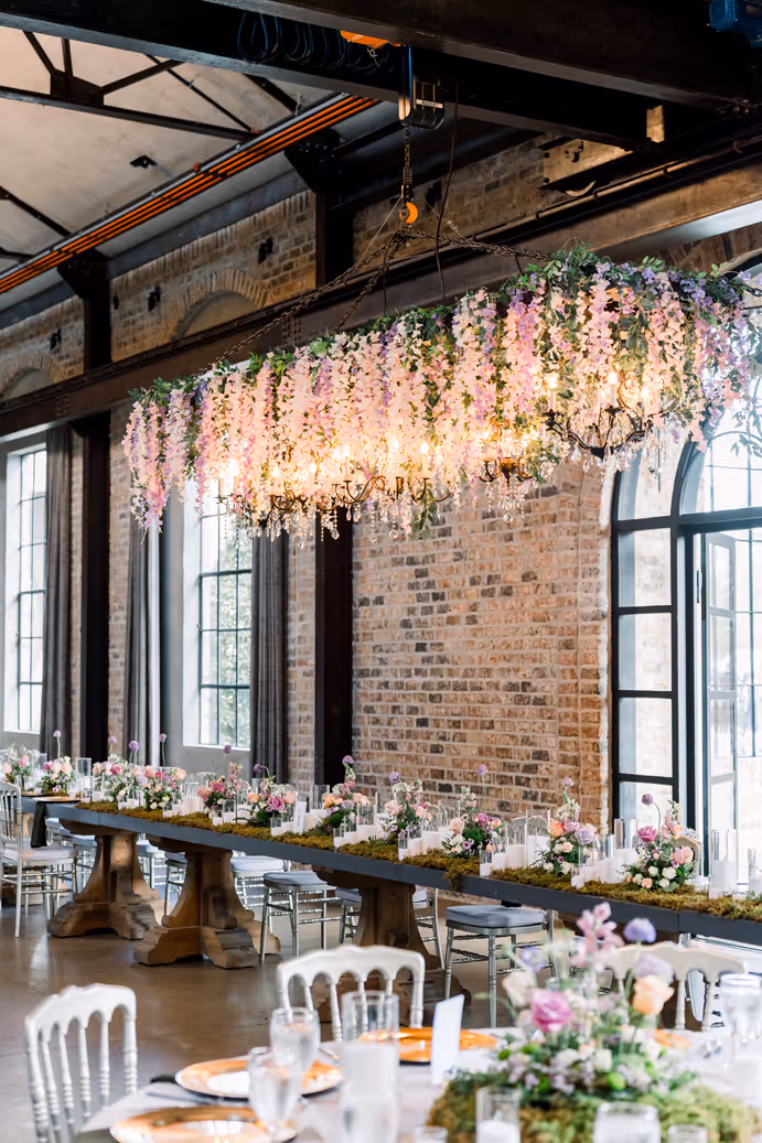 Indoor dining setup with a long wooden table decorated with flowers and candles beneath a chandelier covered in cascading pink and white flowers.