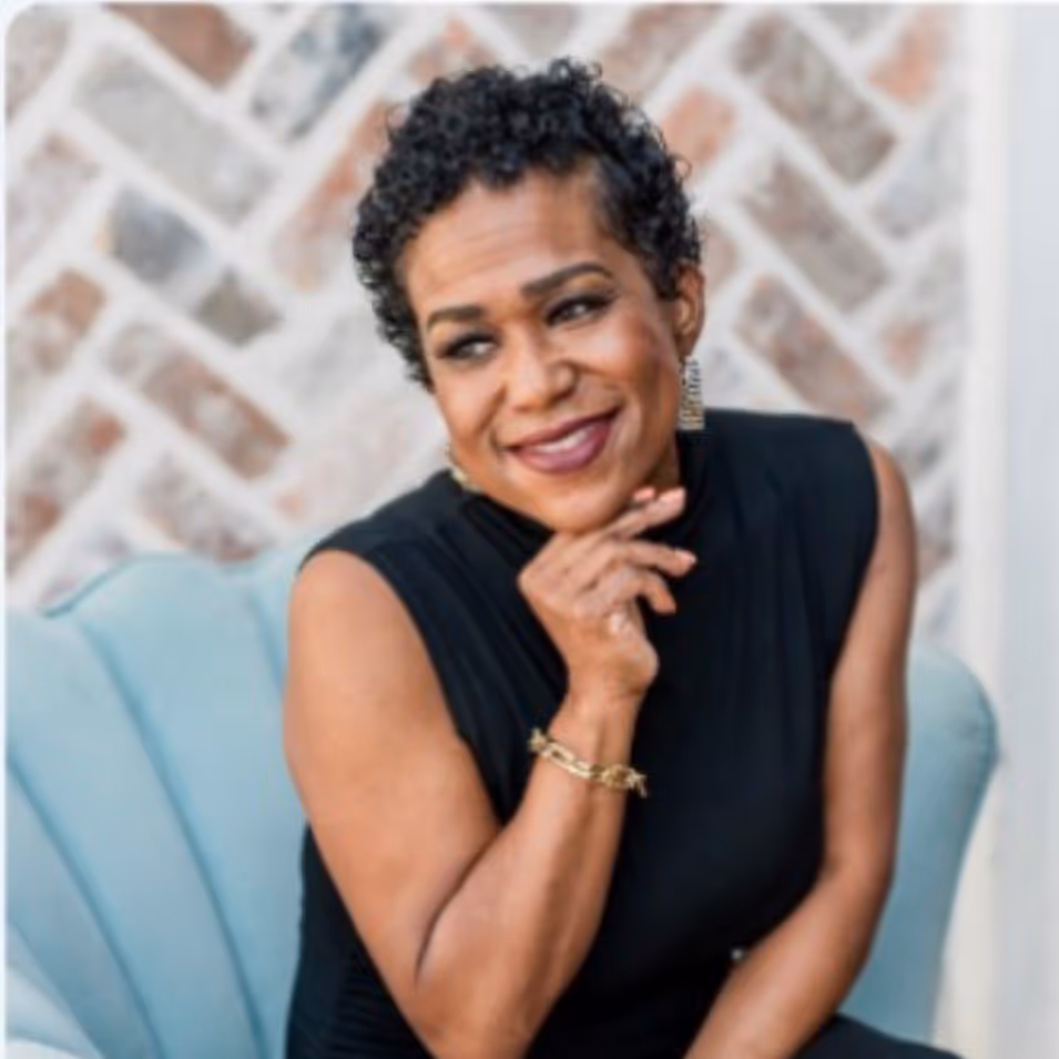 Smiling woman with short curly hair wearing a sleeveless black top, seated on a light blue chair against a blurred brick wall.