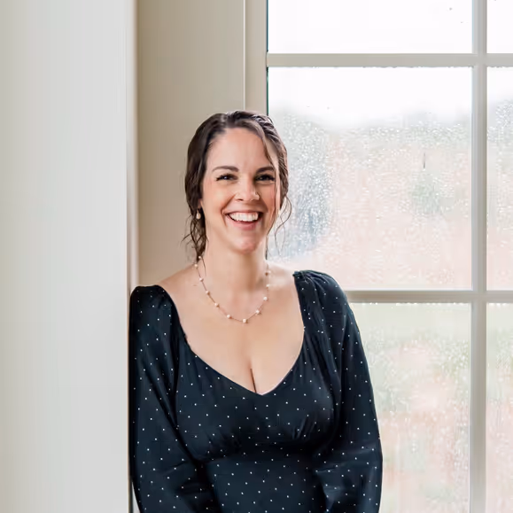 Smiling woman with braided hair wearing a black dress with white dots, standing by a window with raindrops.