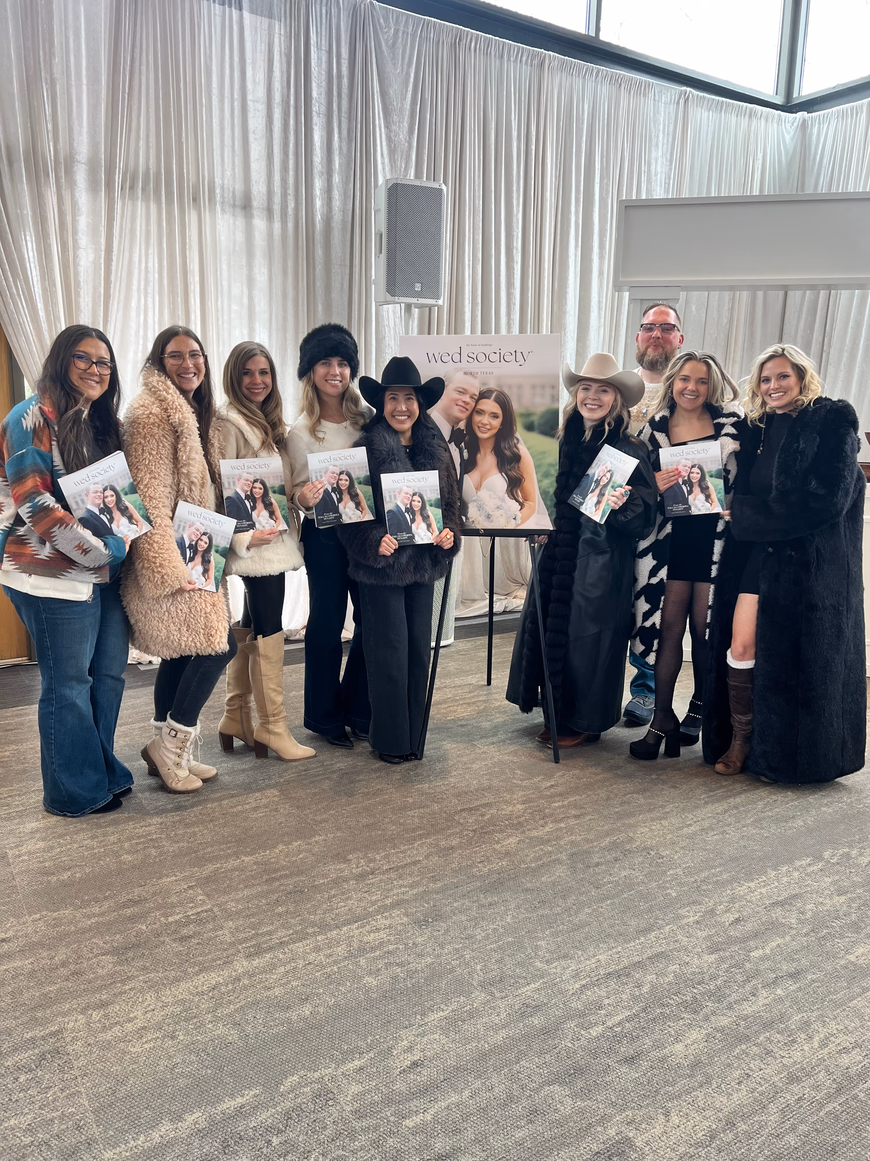 Group of nine people, mostly women dressed in winter clothing, holding Wed Society magazines next to a large Wed Society North Texas poster.