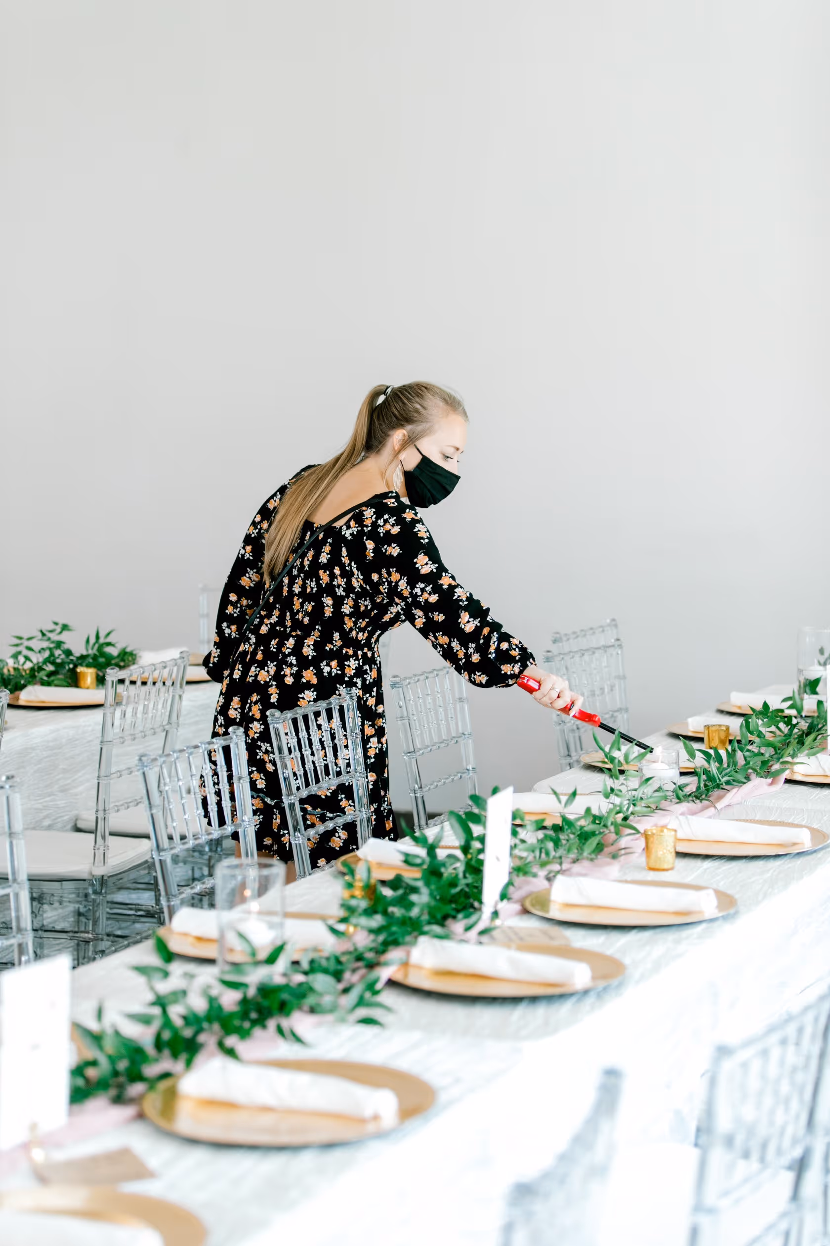 Woman in a black floral dress lighting candles on a decorated table with greenery and gold plates.