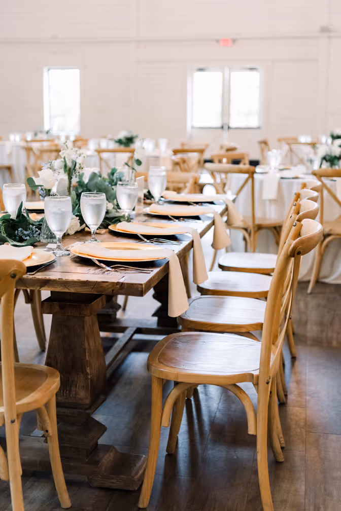 Wooden banquet table set with plates, silverware, water glasses, and floral centerpieces in a bright event space.