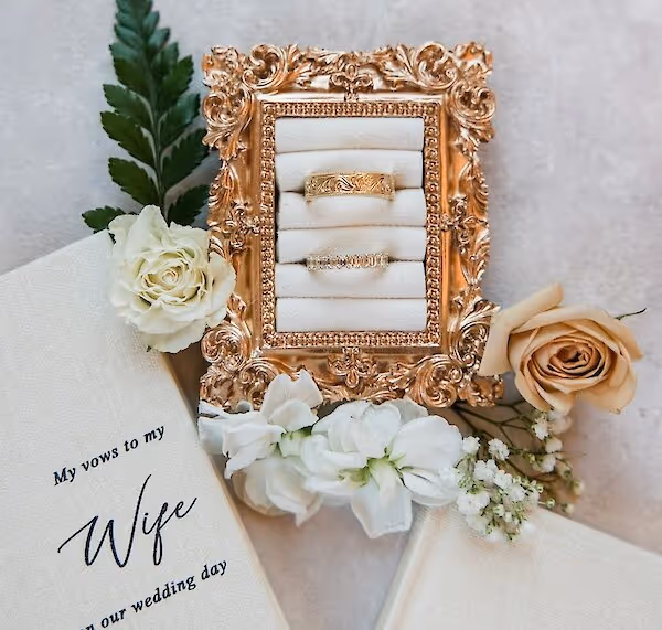 Ornate gold frame holding two wedding rings surrounded by white and peach roses, greenery, and a vow booklet titled 'My vows to my Wife on our wedding day'.