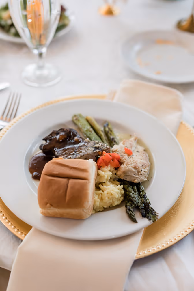 Plate with grilled asparagus, chicken breast with diced tomatoes, braised meat in sauce, yellow rice, and a bread roll on a white napkin.