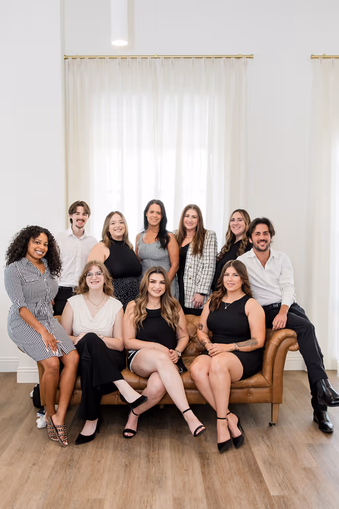 Group of nine young adults, six women and three men, posing together indoors on and around a brown leather couch in front of large windows with white curtains.