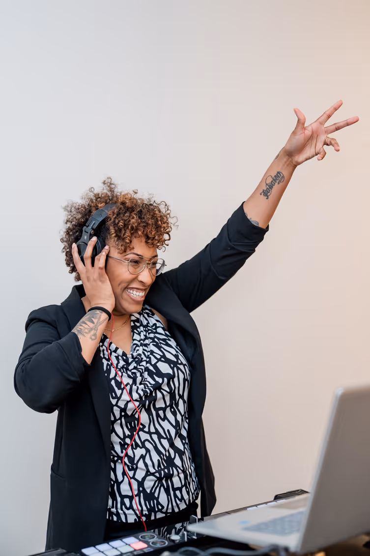 Smiling DJ with curly hair and glasses wearing headphones, raising one arm while mixing music on a laptop and DJ controller.