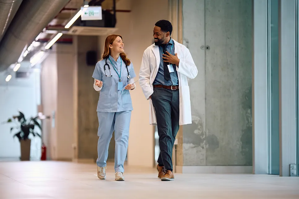 A female nurse and a male doctor walking and smiling while talking in a hospital corridor.