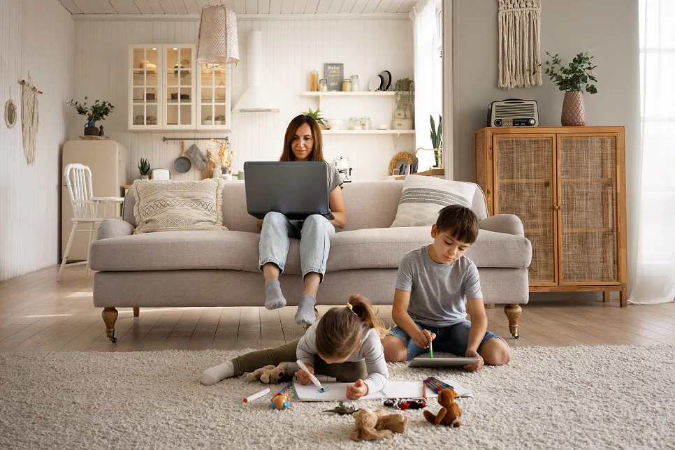 Woman sitting on a beige sofa using a laptop while two children sit on a carpet drawing and coloring in a cozy living room.