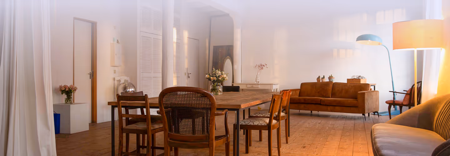 Gemütlich eingerichtetes Wohnzimmer mit Holztisch und verschiedenen Stühlen, braunem Sofa und warmem Bodentischlampenlicht.