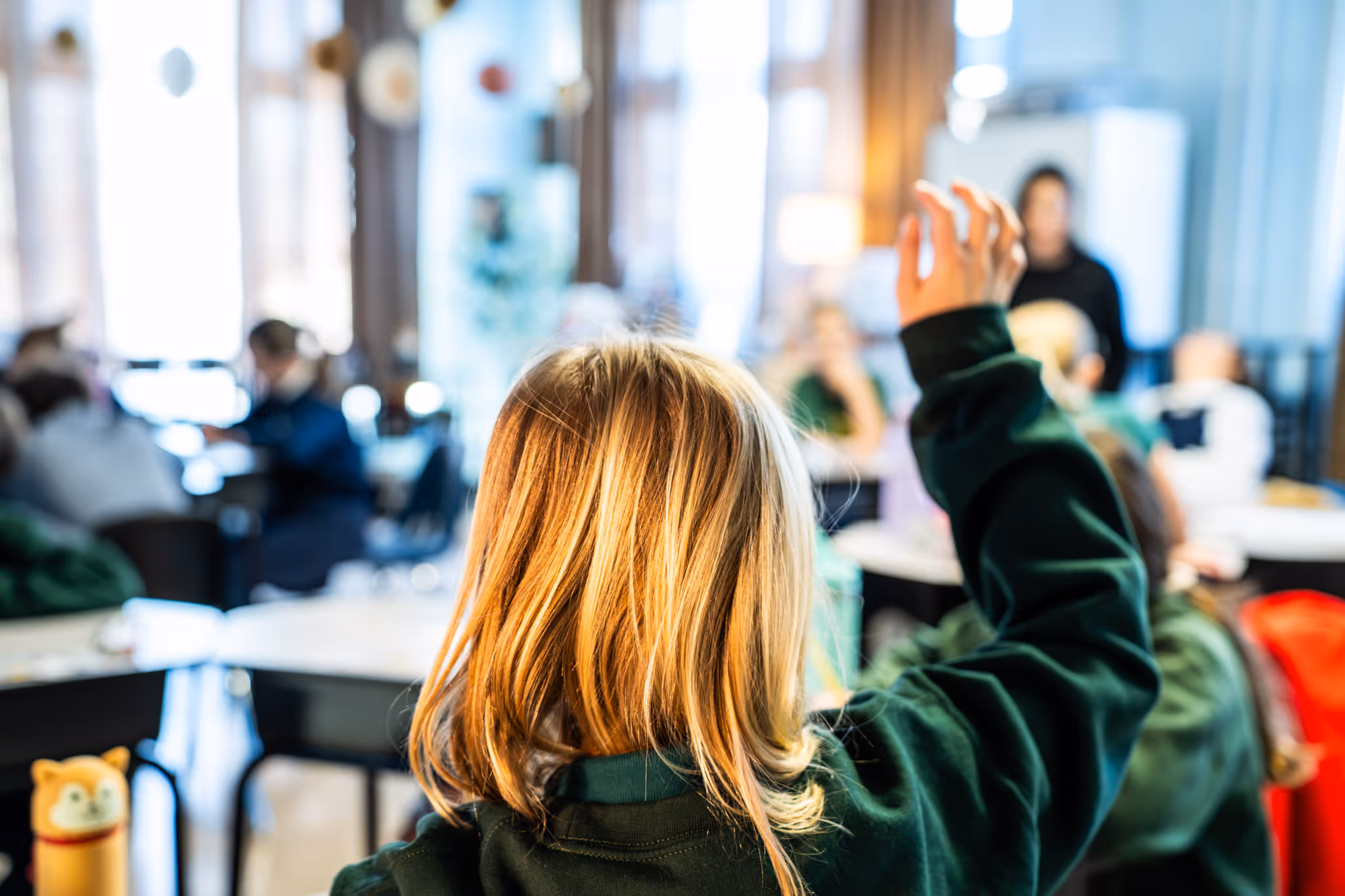 Student with blonde hair raising hand in a classroom with other children and a teacher.