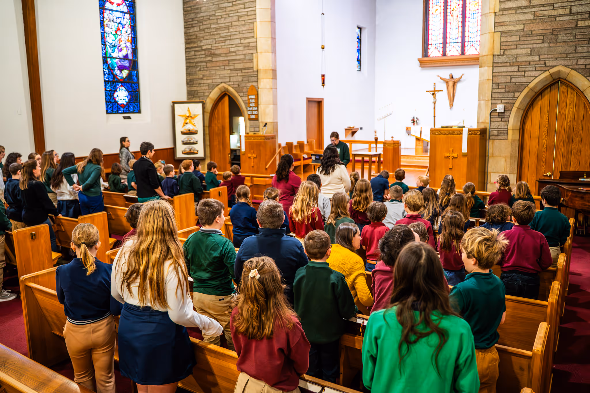 Children and adults standing in wooden pews inside a church with stained glass windows and a pastor leading at the altar.