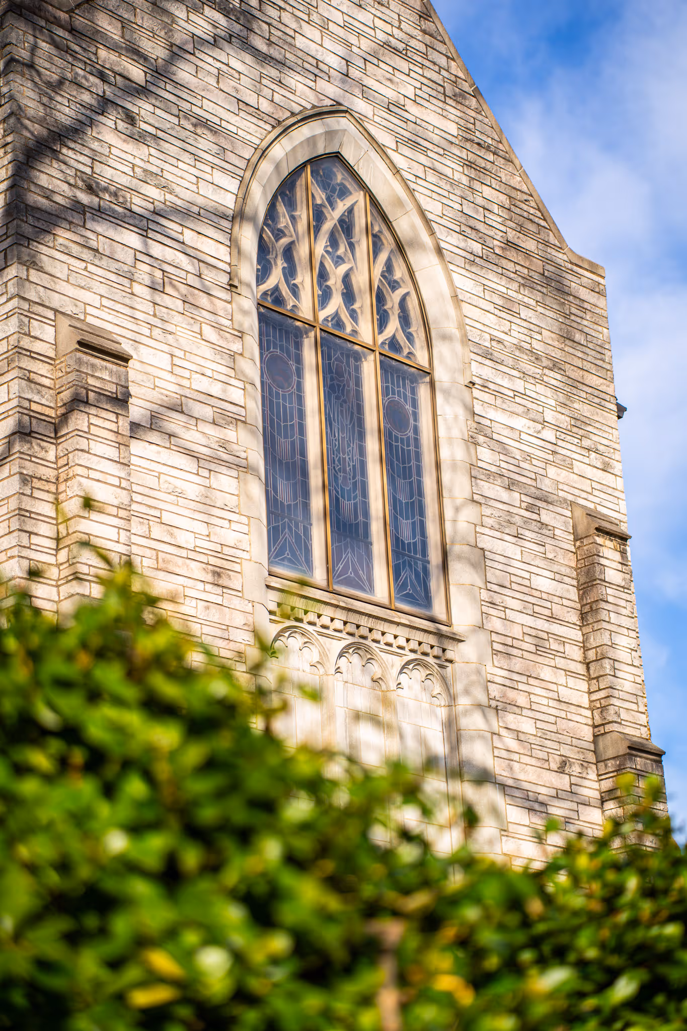 Close-up of a church wall with a tall arched stained glass window and green foliage in the foreground.