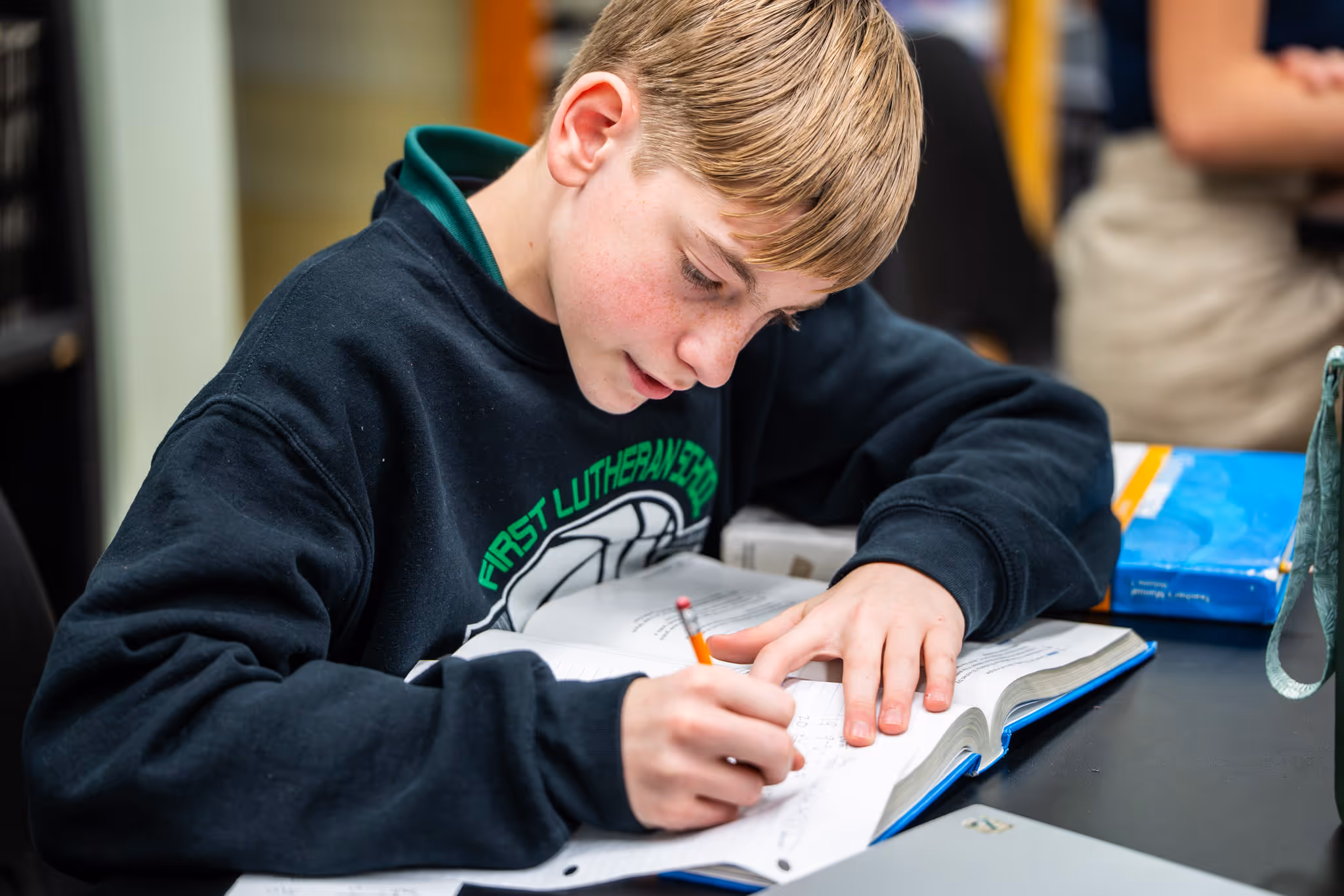 Boy in a black First Lutheran School sweatshirt focused on writing in a notebook with a pencil at a desk.